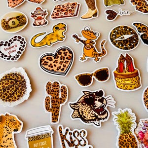 Könnte beinhalten: Eine Sammlung von gestanzten Aufklebern mit verschiedenen Leopardenmustern. Die Motive umfassen einen Basketball, einen Kaktus, einen Kaffeebecher mit dem Text 'BUT FIRST, coffee', einen Leopardenkopf, eine Ananas, eine Sonnenbrille und ein Herz.