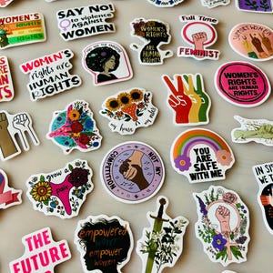 Könnte beinhalten: Eine Sammlung verschiedener Vinyl-Aufkleber mit unterschiedlichen Motiven, darunter erhobene Fäuste, Blumenmuster und Text-Slogans wie "The Future Is Female", "Women's Rights Are Human Rights" und "Feminist", die in einem gitterartigen Muster angeordnet sind.