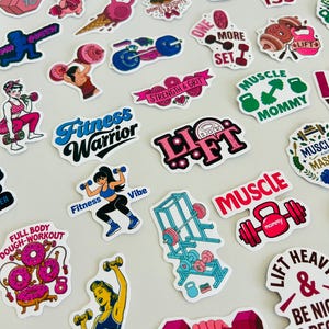 Könnte beinhalten: Eine Sammlung bunter Vinyl-Aufkleber mit Fitness-Illustrationen, darunter Hanteln, Kettlebells, Langhanteln, Hantelständer und motivierende Texte wie Fitness Warrior, Lift und Muscle Mommy, angeordnet auf einer glatten Oberfläche.
