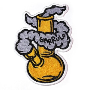 Puede incluir: Parche bordado que representa un bong amarillo con humo gris saliendo de la parte superior. La palabra "GARRULO" está bordada en blanco sobre el humo. El parche tiene un borde blanco.