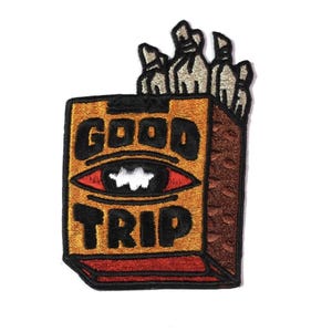 Può includere: Toppa ricamata con le parole "GOOD TRIP" e un disegno a occhio. La toppa ha uno schema di colori oro, marrone, rosso e nero. La parte superiore della scatola ha diversi oggetti color argento. Per abbigliamento.