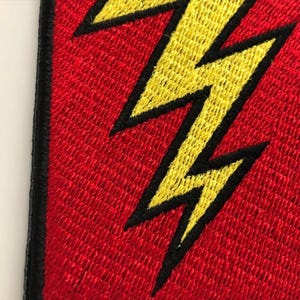 Pode incluir: Um emblema de tecido com um design de raio amarelo brilhante com um contorno preto espesso, sobre um fundo vermelho vibrante. A pe&ccedil;a &eacute; finalizada com uma borda bordada preta densa ao longo do lado vertical esquerdo do objeto.