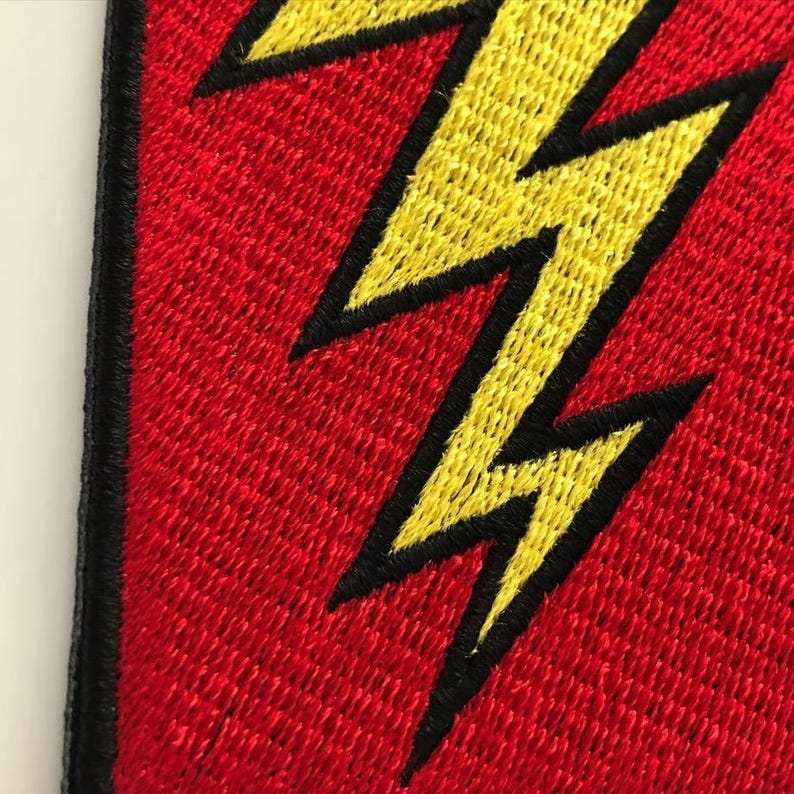 Pode incluir: Um emblema de tecido com um design de raio amarelo brilhante com um contorno preto espesso, sobre um fundo vermelho vibrante. A pe&ccedil;a &eacute; finalizada com uma borda bordada preta densa ao longo do lado vertical esquerdo do objeto.