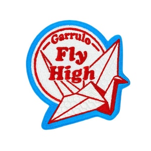 May include: A circular embroidered patch with a bright blue border. The center features a white background with red text that reads Garrulo Fly High, accompanied by a white paper crane graphic with red outlines and accents.