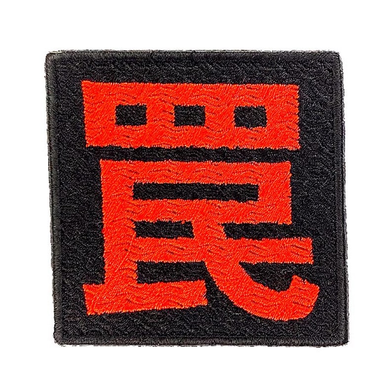 Puede incluir: Un parche bordado de forma cuadrada que presenta un fondo negro con un car&aacute;cter kanji japon&eacute;s de color rojo brillante centrado en la superficie. El bordado tiene una apariencia texturizada y en relieve con un borde definido.