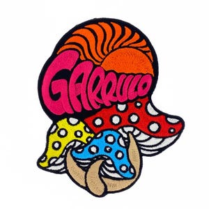 May include: Embroidered patch featuring a colorful design of mushrooms and the word "Garrulo" in pink. The patch includes a red mushroom with white spots, a yellow mushroom, and a blue mushroom, all with a black border.