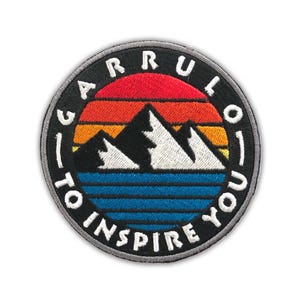 May include: A circular embroidered patch featuring a mountain landscape with white peaks against a sunset of red, orange, and yellow stripes above a blue lake. The black border contains the white embroidered text GARRULO TO INSPIRE YOU.