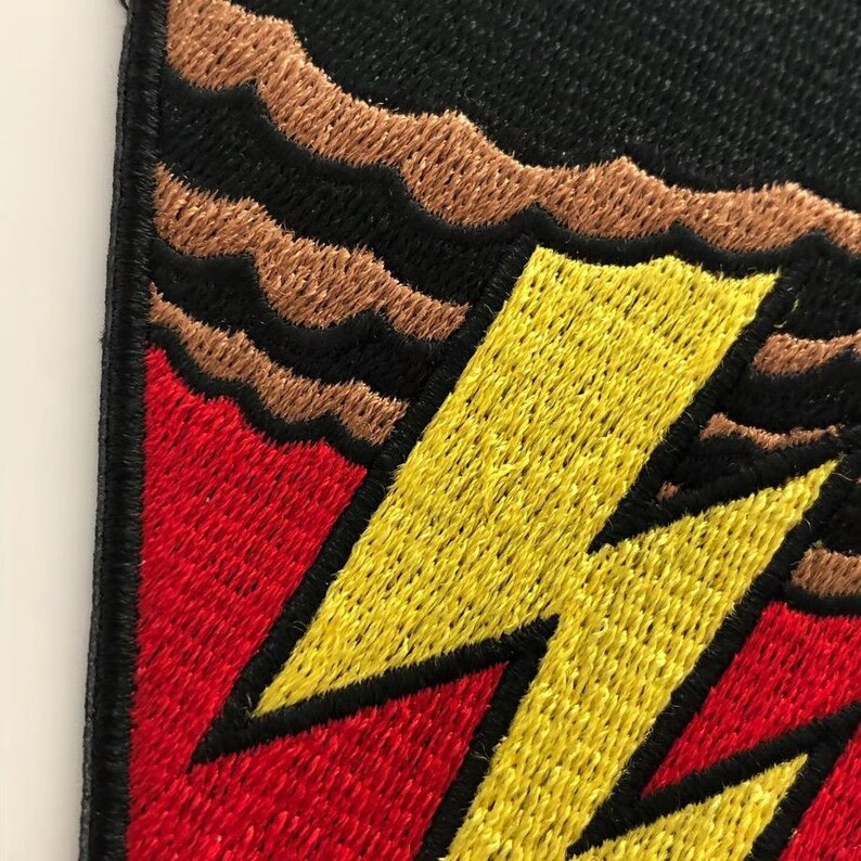 Pode incluir: Um emblema bordado com um raio amarelo brilhante sobre um fundo vermelho vibrante. O design inclui faixas horizontais castanhas onduladas na parte superior, tudo contornado por uma espessa borda preta costurada sobre uma base preta.