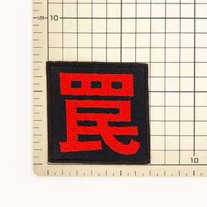Puede incluir: Un parche bordado de forma cuadrada con fondo negro y un car&aacute;cter kanji japon&eacute;s de color rojo intenso centrado en la tela. El parche est&aacute; colocado sobre una alfombrilla de cuadr&iacute;cula con marcas que indican un tama&ntilde;o de unos 4 cent&iacute;metros.