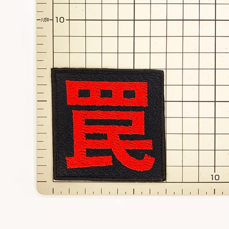 Puede incluir: Un parche bordado de forma cuadrada con fondo negro y un car&aacute;cter kanji japon&eacute;s de color rojo intenso centrado en la tela. El parche est&aacute; colocado sobre una alfombrilla de cuadr&iacute;cula con marcas que indican un tama&ntilde;o de unos 4 cent&iacute;metros.