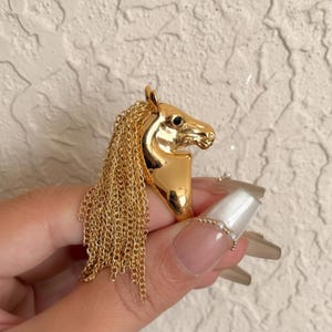 May include: A gold-colored ring featuring a horse head design with a flowing chain mane. The horse's eye is a small black gem. The ring is a statement piece, likely made of metal, and has a polished finish.