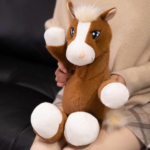 Puede incluir: Un juguete de peluche de caballo marrón y blanco. El juguete tiene una mancha blanca en la cara, cascos blancos y una melena y cola blancas y esponjosas. El caballo está siendo sostenido por una persona.