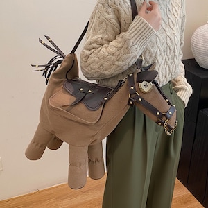 Op de afbeelding: Een schoudertas in de vorm van een paard. De tas is voornamelijk bruin met bruine zadel- en teugeldetails. De paardenkop heeft een goudkleurige ketting en een decoratief kruis.