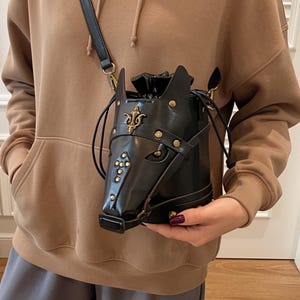 Può includere: Borsa a secchiello nera a forma di testa di cavallo, con chiusura a coulisse e tracolla. La borsa presenta borchie e dettagli decorativi color oro. Il design include un volto di cavallo dettagliato.