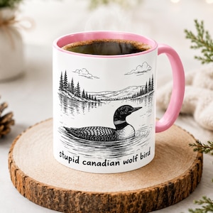 May include: A white ceramic mug with a pink interior and handle, featuring a black and white line art illustration of a loon swimming on a lake with pine trees and clouds in the background, and the text "stupid canadian wolf bird" printed below.