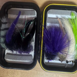 May include: Two open black fly fishing tackle boxes with yellow edging, filled with colourful fishing flies. The flies feature purple, black, white, and green feathers and synthetic materials. The boxes are open, revealing the interior compartments and fly arrangements.