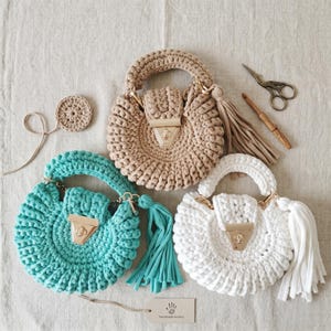Puede incluir: Tres bolsos redondos de ganchillo en beige, turquesa y blanco, cada uno con asa, cierre de metal dorado y borla de tela a juego. Se incluyen una pequeña pieza de ganchillo, unas tijeras, una herramienta de madera y una etiqueta.