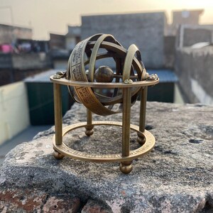 Op de afbeelding: Een messing armillairsfeer, een model van een hemelglobe, staat op een stenen oppervlak. De bol heeft concentrische ringen met gegraveerde tekst en een centrale bol. De basis heeft een cirkelvormig ontwerp met cijfers en tekst, ondersteund door vier poten.