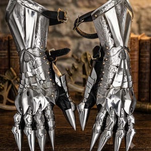 May include: A pair of polished silver metal gauntlets featuring intricate floral engravings, articulated finger plates with pointed tips, and black leather straps with brass-toned buckles for secure fastening around the wrist and forearm.