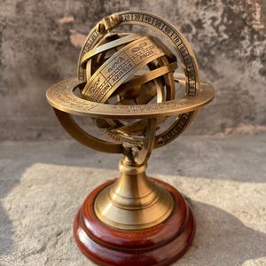 Puede incluir: Una esfera armilar de latón con símbolos del zodiaco, montada sobre una base de madera. La esfera presenta anillos con marcas de grados y los signos del zodiaco Capricornio, Acuario y Piscis. La base es de color marrón rojizo oscuro.