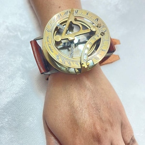 Op de afbeelding: Een vintage messing zonnewijzerhorloge met een bruine leren band. De wijzerplaat heeft Romeinse cijfers en de tekst "West London". Het horloge wordt om de pols gedragen.