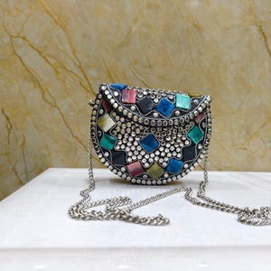 Op de afbeelding: Een zilverkleurige, halvemaanvormige handtas met een kettingriem. De tas is versierd met kleurrijke, vierkante edelstenen en kleine, zilveren studs. De edelstenen zijn in tinten blauw, roze, groen en geel. De tas staat op een wit oppervlak.
