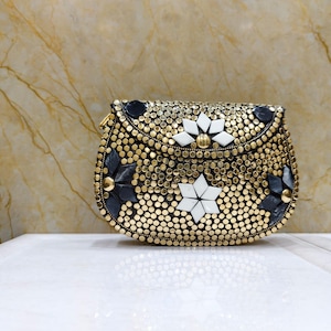 Op de afbeelding: Een kleine, goudkleurige handtas met een afgeronde vorm. De tas is bedekt met kleine, ronde studs en heeft decoratieve zwarte en witte bloemaccenten. Hij heeft een goudkleurige ketting en een sluiting met een sluiting.