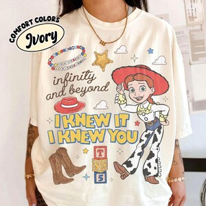 May include: An ivory t-shirt featuring a graphic print of a cowgirl character, a red cowboy hat, brown cowboy boots, a beaded bracelet, a star, and alphabet blocks. The text reads: You've got a friend in me, infinity and beyond, I knew it, I knew you.