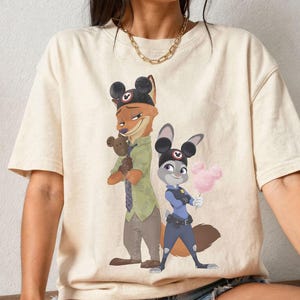 Pu&ograve; includere: T-shirt color crema con un'illustrazione di due personaggi di Zootropolis. Uno &egrave; una volpe con un cappello con orecchie da topo e un orsacchiotto. L'altro &egrave; un coniglio in uniforme da poliziotto, con zucchero filato rosa.