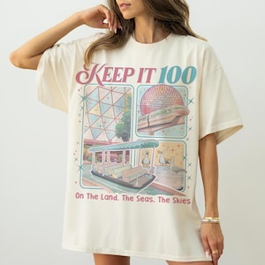 May include: Cream-colored oversized t-shirt with a colorful graphic design. The design features illustrations of a boat, a geodesic sphere, and other theme park elements, with the text "KEEP IT 100" and "On The Land. The Seas. The Skies."