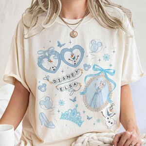 May include: Cream-colored t-shirt featuring a Disney Elsa design. The design includes images of Elsa, Olaf, and other Frozen-themed elements in shades of blue. The text "Disney Elsa" is also present.