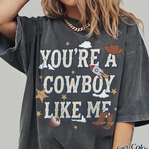 May include: A charcoal gray t-shirt featuring the text "YOU'RE A COWBOY LIKE ME" in a distressed font. The graphic includes a brown cowboy hat, a rocket ship, a sheriff star badge labeled "TAYLOR", a red and white ball, and brown cowboy boots.