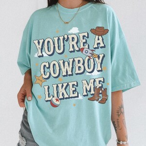 May include: A light blue t-shirt featuring the text "YOU'RE A COWBOY LIKE ME" in a dark blue font. The design includes a brown cowboy hat, a rocket ship, a yellow sheriff star badge labeled "TAYLOR", a red and white ball, and brown cowboy boots.
