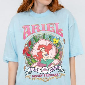 May include: Pale blue t-shirt with a graphic of Ariel from The Little Mermaid, seated in a seashell. The design includes the word "ARIEL" in pink, the year "1989", and the words "Disney Princess".