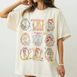 May include: Cream-colored t-shirt featuring nine framed portraits of animated princesses. Each portrait is surrounded by a decorative gold frame with floral and bow accents. The portraits include various princesses in their signature attire.