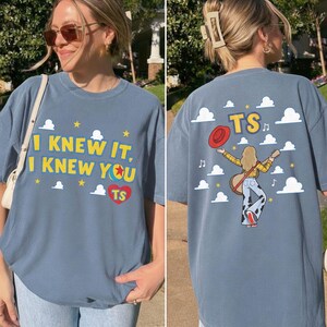 May include: A blue t-shirt featuring a graphic design. The front displays the text I KNEW IT, I KNEW YOU with a ball icon and a red heart containing TS. The back shows a person with a guitar, a red hat, and the text TS amidst white clouds.
