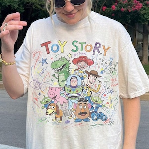 K&ouml;nnte beinhalten: Cremefarbenes T-Shirt mit dem Schriftzug "TOY STORY" in bunten Buchstaben &uuml;ber einer Grafik von Figuren aus dem Animationsfilm. Das Design umfasst Woody, Buzz Lightyear und andere Charaktere mit bunten Kritzeleien.