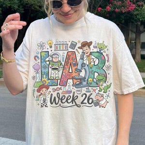 Op de afbeelding: Crèmekleurig T-shirt met het woord "LAB" in kleurrijke letters, met cartoonfiguren en wetenschappelijke illustraties. De tekst "Week 26" staat onder het woord "LAB".