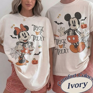 May include: Two ivory t-shirts featuring Halloween-themed graphics of Mickey and Minnie Mouse in skeleton costumes. The designs include the text "Trick or Treat," spiderwebs, bats, pumpkins, and candy corn, printed in black, orange, and white colors.