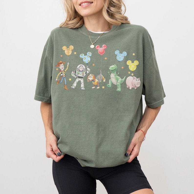 May include: Green t-shirt featuring a graphic of Toy Story characters and Mickey Mouse-shaped balloons. The design includes Woody, Buzz Lightyear, Rex, Hamm, and Slinky Dog. The shirt is a relaxed fit and has short sleeves.