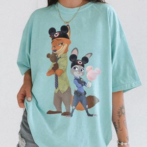 Pu&ograve; includere: Maglietta azzurra con un'illustrazione di Nick Wilde e Judy Hopps di Zootropolis, entrambi con orecchie di Topolino. Nick, una volpe, tiene un orsacchiotto, e Judy, una coniglia, tiene zucchero filato rosa a forma di Topolino.