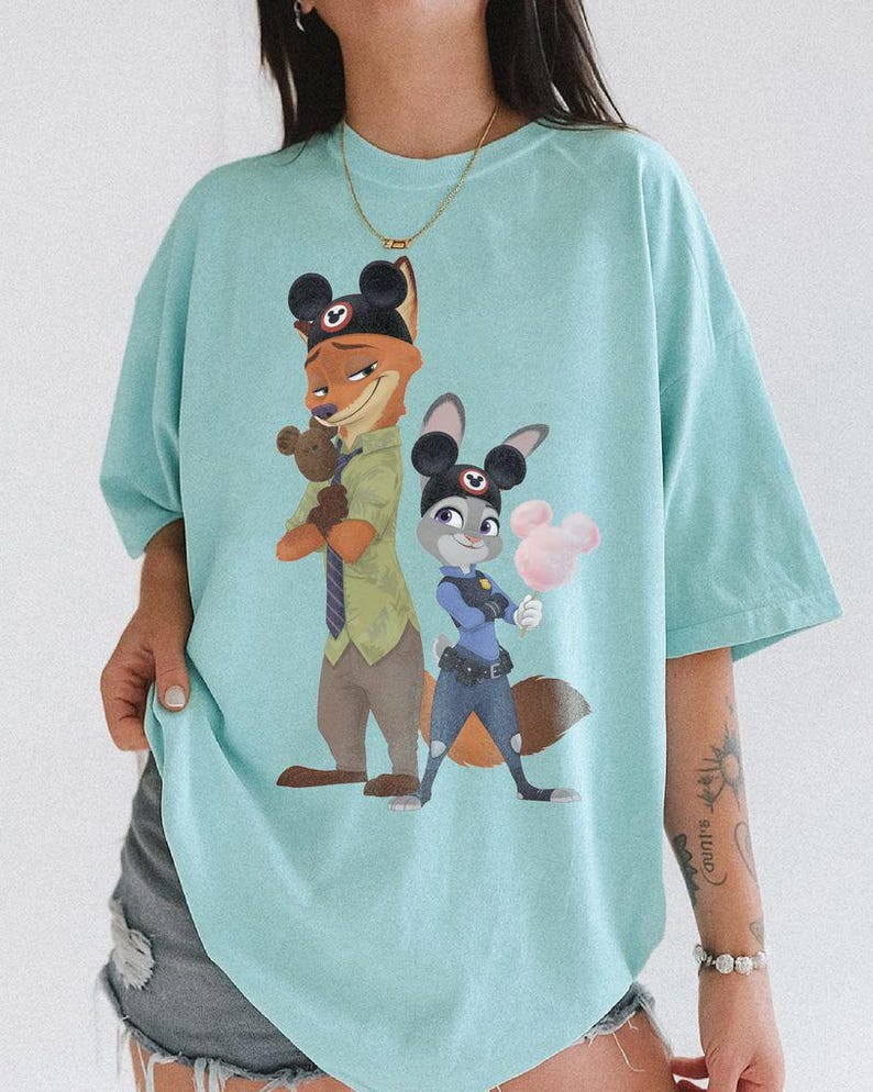 Pu&ograve; includere: Maglietta azzurra con un'illustrazione di Nick Wilde e Judy Hopps di Zootropolis, entrambi con orecchie di Topolino. Nick, una volpe, tiene un orsacchiotto, e Judy, una coniglia, tiene zucchero filato rosa a forma di Topolino.