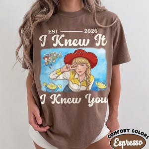 May include: A brown short-sleeve t-shirt featuring a graphic print of a blonde character in a red cowboy hat making a peace sign, surrounded by animated characters. The text reads EST 2026, I Knew It, and I Knew You in a cream-colored font.