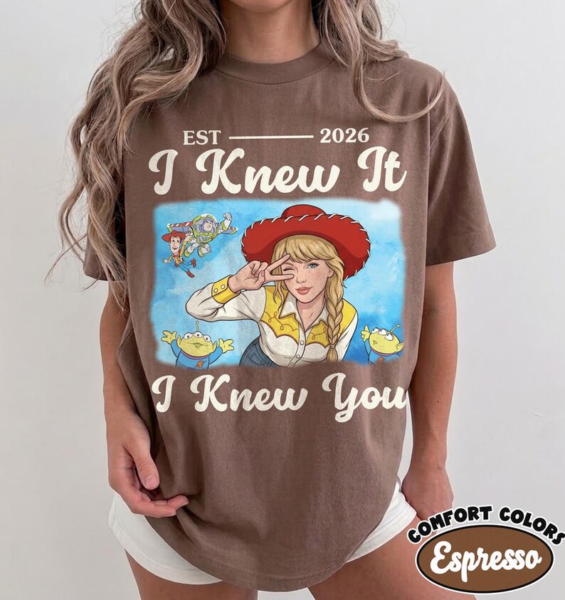 May include: A brown short-sleeve t-shirt featuring a graphic print of a blonde character in a red cowboy hat making a peace sign, surrounded by animated characters. The text reads EST 2026, I Knew It, and I Knew You in a cream-colored font.