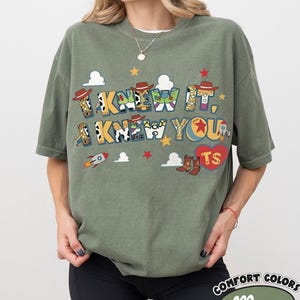 May include: An olive green short-sleeve t-shirt featuring the text I KNEW IT, I KNEW YOU in a colorful, patterned font. The design includes small illustrations of cowboy hats, rockets, clouds, stars, and a red heart with the letters TS inside.