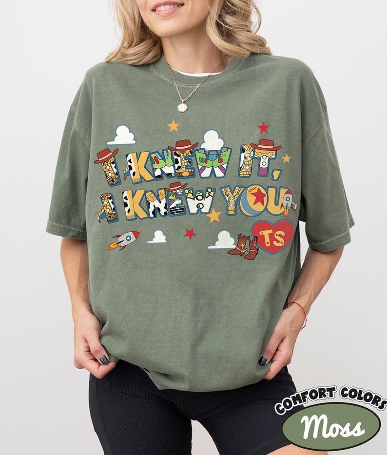 May include: An olive green short-sleeve t-shirt featuring the text I KNEW IT, I KNEW YOU in a colorful, patterned font. The design includes small illustrations of cowboy hats, rockets, clouds, stars, and a red heart with the letters TS inside.