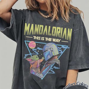 Może przedstawiać: Ciemnoszary t-shirt z nadrukiem graficznym przedstawiającym Mandalorianina i Grogu. Napis głosi MANDALORIAN oraz THIS IS THE WAY. Wz&oacute;r zawiera niebieski tr&oacute;jkąt, r&oacute;żową planetę i gwiazdy. Koszulka ma wyblakły, stylizowany na vintage wygląd.
