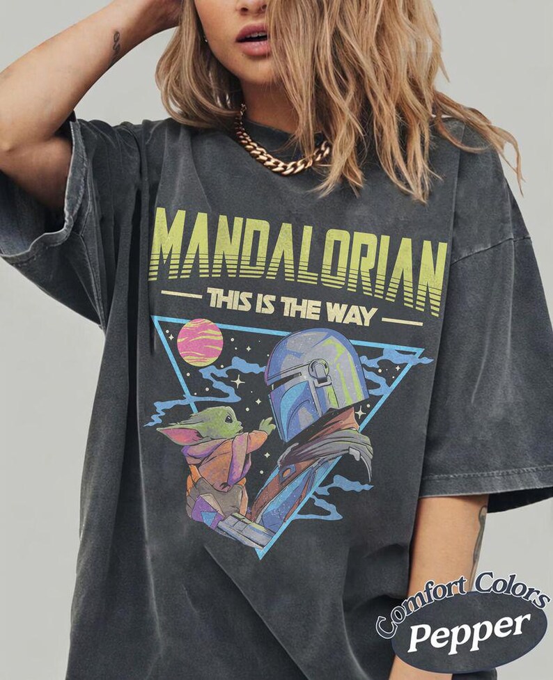 Może przedstawiać: Ciemnoszary t-shirt z nadrukiem graficznym przedstawiającym Mandalorianina i Grogu. Napis głosi MANDALORIAN oraz THIS IS THE WAY. Wz&oacute;r zawiera niebieski tr&oacute;jkąt, r&oacute;żową planetę i gwiazdy. Koszulka ma wyblakły, stylizowany na vintage wygląd.