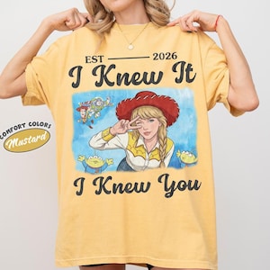 May include: A mustard yellow t-shirt featuring a graphic print with the text EST 2026 I Knew It I Knew You. The illustration shows a blonde character in a red cowboy hat, Woody, Buzz Lightyear, and two green aliens on a blue watercolor background.