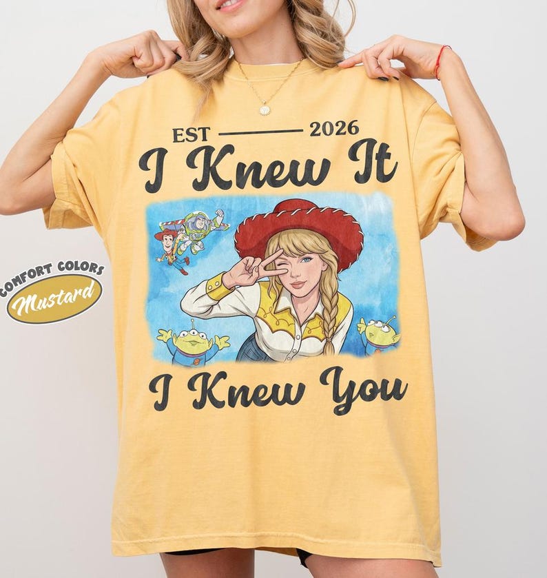May include: A mustard yellow t-shirt featuring a graphic print with the text EST 2026 I Knew It I Knew You. The illustration shows a blonde character in a red cowboy hat, Woody, Buzz Lightyear, and two green aliens on a blue watercolor background.